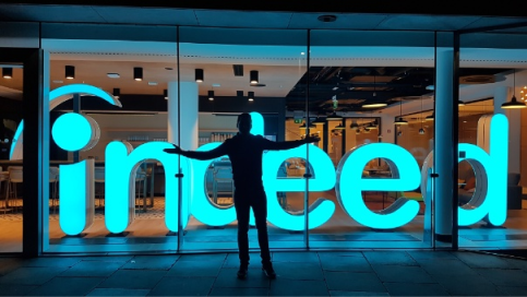 Located
        in front of the Indeed headquarters, there is a silhouette of a person
        with outstretched hands standing in front of a large Indeed neon
        sign.